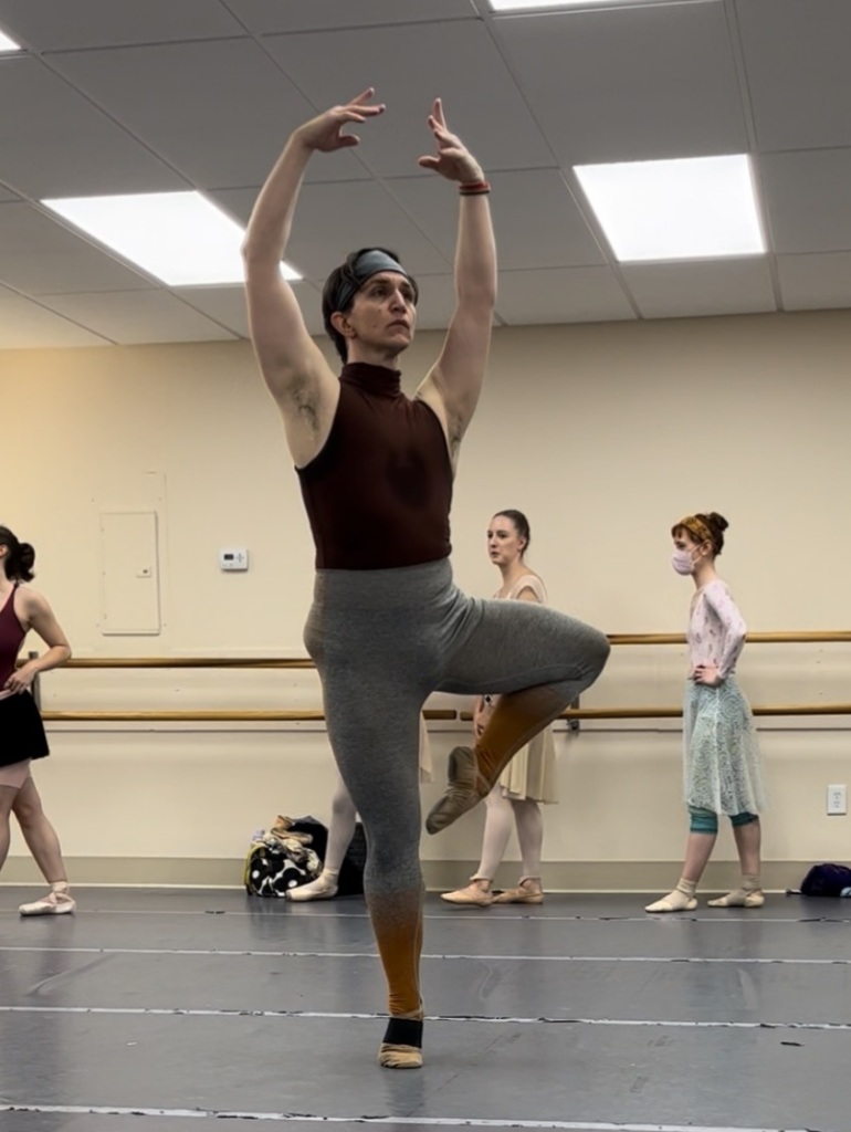 The author executing a pirouette in retire in grey-to-orange ombre tights and a brown halter-neck leotard with other dancers in the background.
